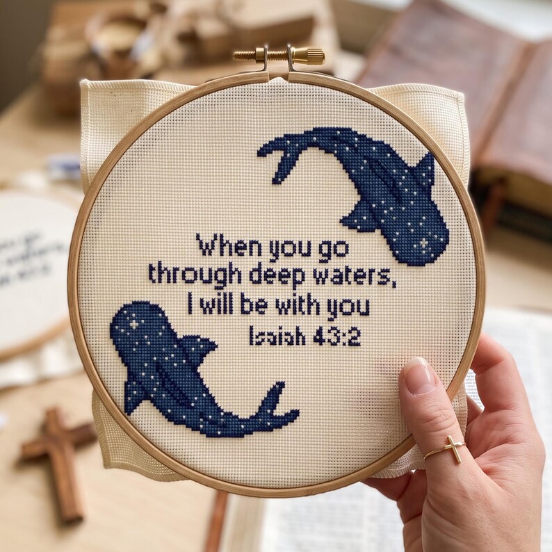 May include: A circular wooden embroidery hoop containing cream-coloured fabric with a cross-stitch design of two dark blue whale sharks and the text: When you go through deep waters, I will be with you Isaiah 43:2, stitched in dark blue thread.