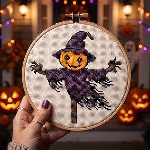 Puede incluir: Un bastidor de bordado circular de madera que contiene un patr&oacute;n de punto de cruz de un espantap&aacute;jaros con una cabeza de calabaza naranja brillante y una t&uacute;nica morada oscura hecha jirones, montado en un poste vertical sobre tela crema.