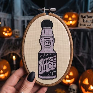 K&ouml;nnte beinhalten: Ein ovaler Stickrahmen aus Holz mit einem Kreuzstichmotiv auf cremefarbenem Stoff. Das Design zeigt eine violette Flasche mit schwarzem Verschluss und dem Text ZOMBIE JUICE in schwarzen Buchstaben sowie ein kleines Schweinegesicht am Flaschenhals.