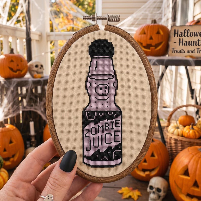 K&ouml;nnte beinhalten: Eine Kreuzstichstickerei in einem ovalen Holzrahmen, die eine hellviolette Flasche mit schwarzem Verschluss und Boden zeigt. Die Flasche hat ein schweineartiges Gesichtsmotiv und den in schwarzem Garn gestickten Text ZOMBIE JUICE auf cremefarbenem Stoff.