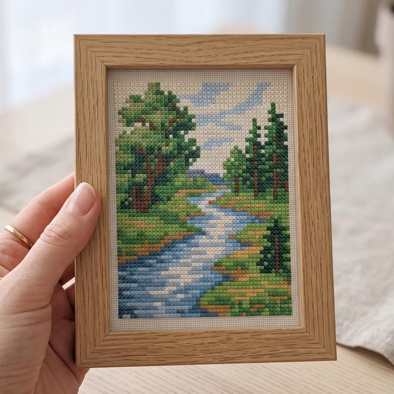 May include: A cross-stitch embroidery depicting a landscape with a winding blue river, green trees, and a light blue sky, framed in a light-colored wooden frame. The needlework features detailed pixelated patterns of nature in various shades of green and blue.