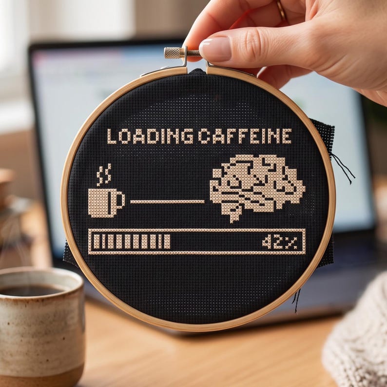 May include: A circular wooden embroidery hoop containing black fabric with a cross-stitch design. The design features the text LOADING CAFFEINE, a coffee mug, a brain, and a progress bar showing 42% completion, all stitched in light beige thread.