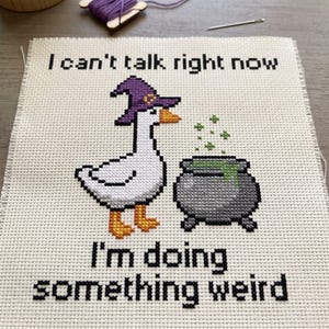 May include: A cross-stitch embroidery on white fabric featuring a white goose wearing a purple witch hat, standing next to a grey cauldron with green bubbling liquid. The text reads: I can't talk right now, I'm doing something weird.