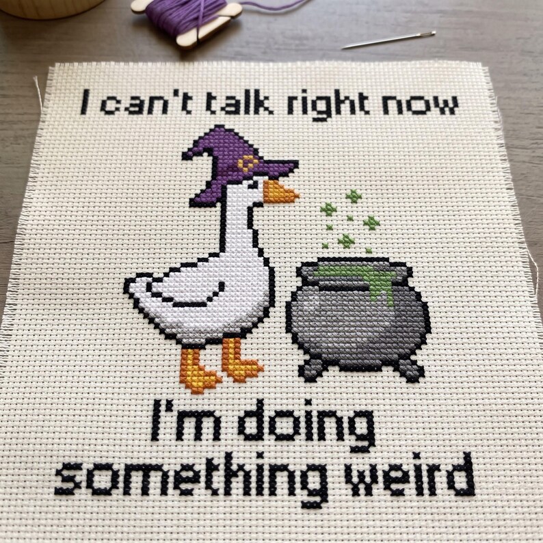 May include: A cross-stitch embroidery on white fabric featuring a white goose wearing a purple witch hat, standing next to a grey cauldron with green bubbling liquid. The text reads: I can't talk right now, I'm doing something weird.