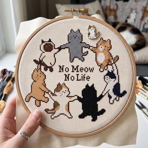 K&ouml;nnte beinhalten: Ein runder Stickrahmen aus Holz mit einem Kreuzstichmuster von sieben Katzen, die sich im Kreis an den Pfoten halten. Der Text "No Meow No Life" ist mittig in braunem Garn gestickt. Eine kleine, katzenf&ouml;rmige Emaille-Anstecknadel ist befestigt.
