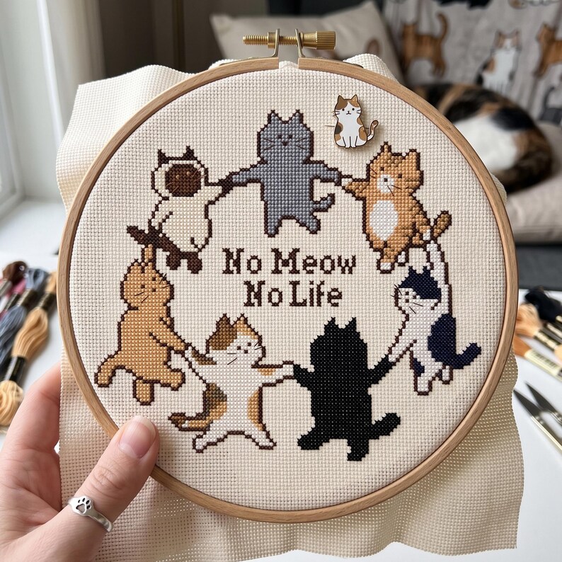 K&ouml;nnte beinhalten: Ein runder Stickrahmen aus Holz mit einem Kreuzstichmuster von sieben Katzen, die sich im Kreis an den Pfoten halten. Der Text "No Meow No Life" ist mittig in braunem Garn gestickt. Eine kleine, katzenf&ouml;rmige Emaille-Anstecknadel ist befestigt.