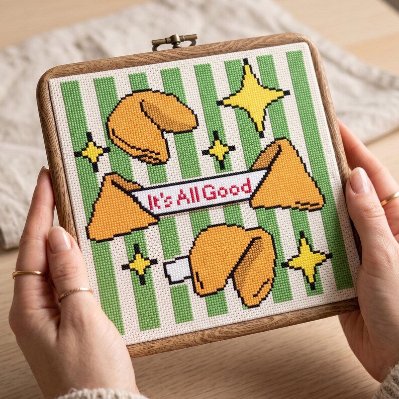 May include: A square cross-stitch embroidery in a wooden frame featuring a green and white striped background. The design includes four yellow fortune cookies and four yellow four-pointed stars. A white banner displays the text It's All Good.