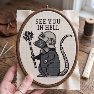 May include: A cross-stitch embroidery in an oval wooden hoop featuring a gray rat wearing a knight's helmet and holding a spiked mace. The text "SEE YOU IN HELL" is stitched in black above the rat on a cream-colored fabric background.