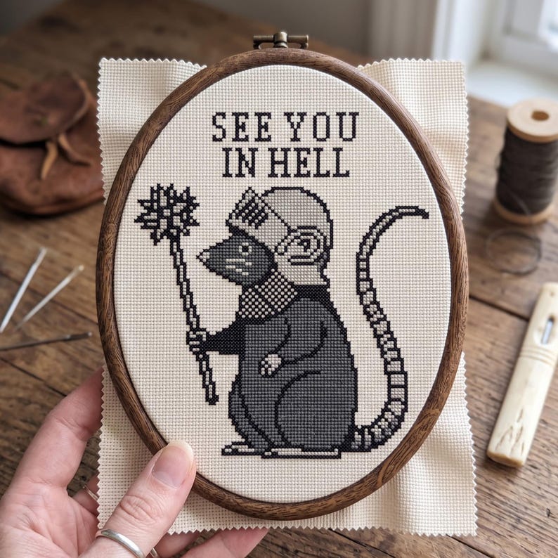 May include: A cross-stitch embroidery in an oval wooden hoop featuring a gray rat wearing a knight's helmet and holding a spiked mace. The text "SEE YOU IN HELL" is stitched in black above the rat on a cream-colored fabric background.