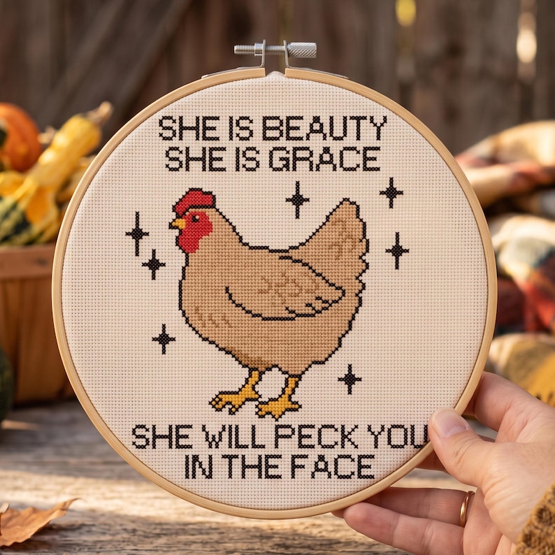 Puede incluir: Un bastidor de bordado redondo de madera con un dise&ntilde;o de punto de cruz de una gallina de color marr&oacute;n claro con cresta roja y patas amarillas. El texto dice: SHE IS BEAUTY SHE IS GRACE SHE WILL PECK YOU IN THE FACE, con estrellas negras.