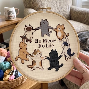 K&ouml;nnte beinhalten: Ein runder Stickrahmen aus Holz mit einem Kreuzstichmotiv auf cremefarbenem Stoff. Das Design zeigt sechs Katzen in verschiedenen Farben, die sich in einem Kreis an den Pfoten halten, mit dem dunkelbraunen Text No Meow No Life in der Mitte.