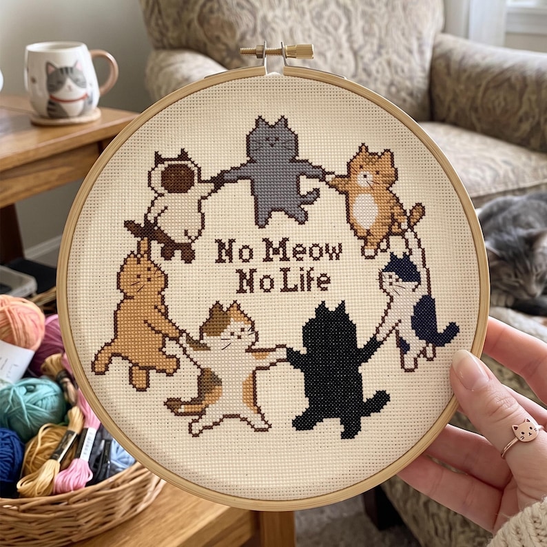 K&ouml;nnte beinhalten: Ein runder Stickrahmen aus Holz mit einem Kreuzstichmotiv auf cremefarbenem Stoff. Das Design zeigt sechs Katzen in verschiedenen Farben, die sich in einem Kreis an den Pfoten halten, mit dem dunkelbraunen Text No Meow No Life in der Mitte.
