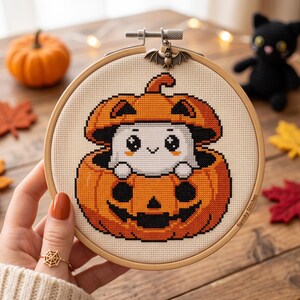 May include: A circular wooden embroidery hoop containing a cross-stitch pattern of a white ghost peeking out from inside an orange jack-o'-lantern pumpkin. A small metal bat charm is attached to the top screw. The text Spooky Stitches is visible.