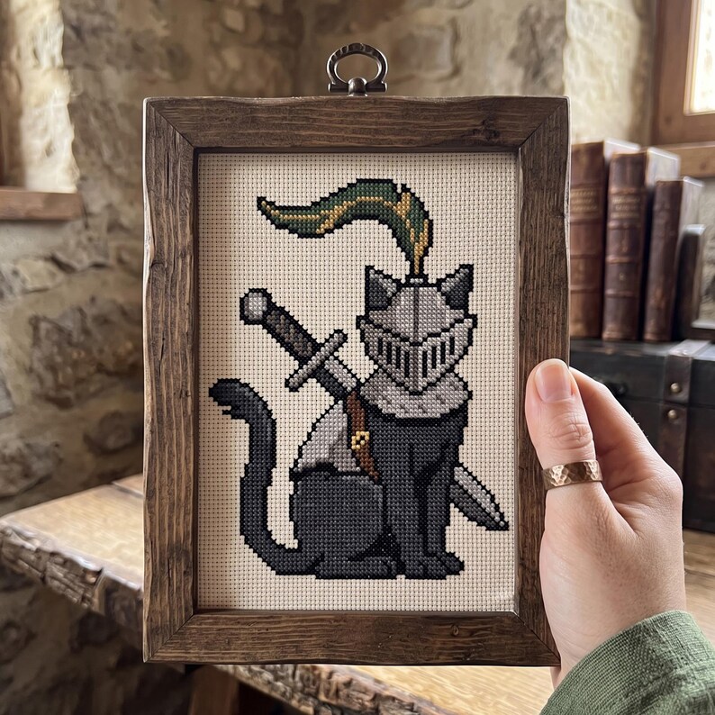 Op de afbeelding: Een kruissteekborduurwerk van een zwarte kat die een zilveren ridderhelm met een groen-gouden pluim draagt, met een zwaard op de rug. Het kunstwerk is ingelijst in een rechthoekige, donkerbruine, rustieke houten lijst met een metalen oogje.