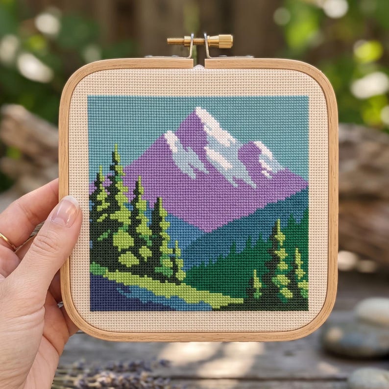 May include: A square wooden embroidery hoop containing a cross-stitch pattern of a mountain landscape. The design features a purple mountain with white snow caps, dark green pine trees, and a light blue sky, all stitched on beige fabric.