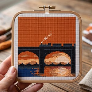 Puede incluir: Un bastidor de bordado cuadrado de madera que contiene una pieza de punto de cruz que representa un tren oscuro sobre un puente contra un cielo naranja vibrante. La escena muestra una locomotora de vapor, arcos y reflejos en el agua.