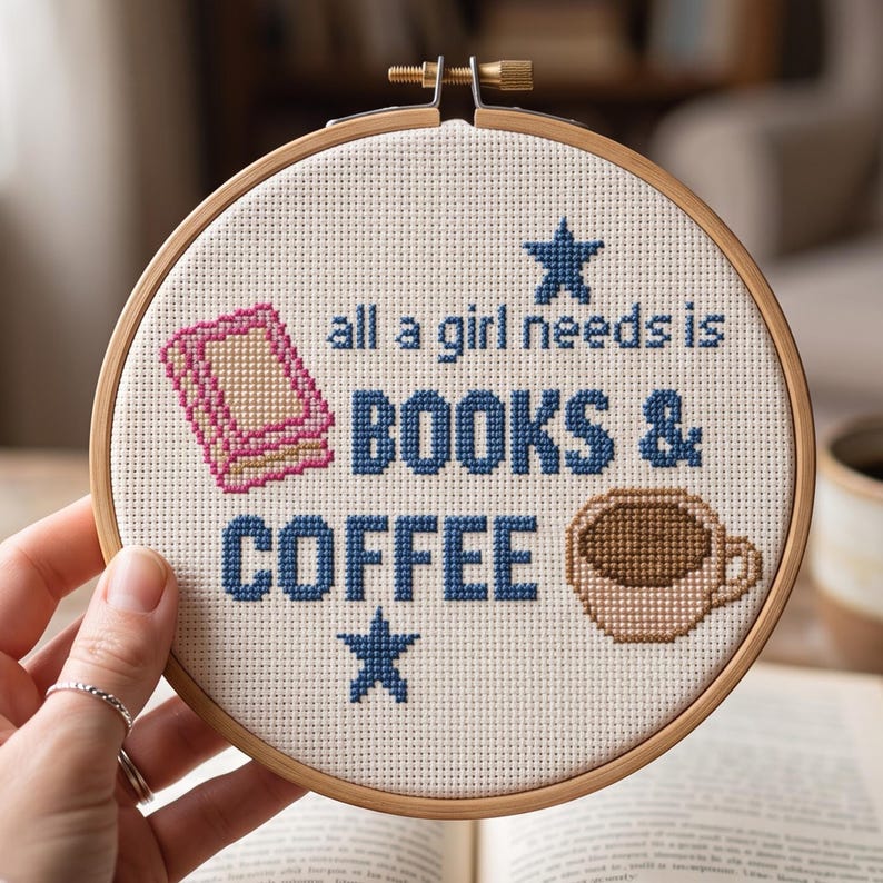May include: A circular wooden embroidery hoop containing white Aida fabric with a cross-stitch design. The text reads all a girl needs is BOOKS & COFFEE in blue. It features a pink book icon, a brown coffee cup, and two blue stars.