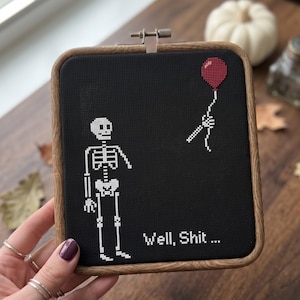 K&ouml;nnte beinhalten: Ein quadratischer Holzstickrahmen mit schwarzem Stoff und einem wei&szlig;en Kreuzstich-Skelett. Dem Skelett fehlt ein Arm, der abgetrennt dar&uuml;ber schwebt und einen roten Luftballon h&auml;lt. Der Text "Well, Shit ..." ist in Wei&szlig; gestickt.
