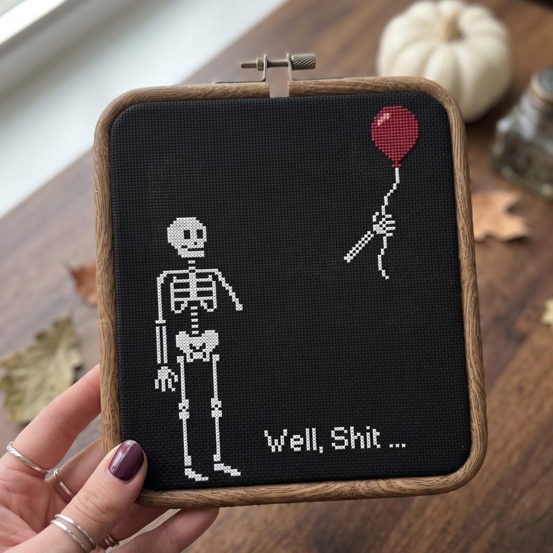 K&ouml;nnte beinhalten: Ein quadratischer Holzstickrahmen mit schwarzem Stoff und einem wei&szlig;en Kreuzstich-Skelett. Dem Skelett fehlt ein Arm, der abgetrennt dar&uuml;ber schwebt und einen roten Luftballon h&auml;lt. Der Text "Well, Shit ..." ist in Wei&szlig; gestickt.