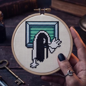 K&ouml;nnte beinhalten: Ein runder Stickrahmen aus Holz mit einem Kreuzstichmotiv auf cremefarbenem Stoff. Das Bild im Pixel-Art-Stil zeigt eine Figur mit langem schwarzem Haar, die aus einem rechteckigen, gr&uuml;n-blauen Fernsehbildschirm mit einer erhobenen Hand hervorkommt.