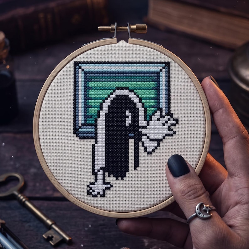 K&ouml;nnte beinhalten: Ein runder Stickrahmen aus Holz mit einem Kreuzstichmotiv auf cremefarbenem Stoff. Das Bild im Pixel-Art-Stil zeigt eine Figur mit langem schwarzem Haar, die aus einem rechteckigen, gr&uuml;n-blauen Fernsehbildschirm mit einer erhobenen Hand hervorkommt.