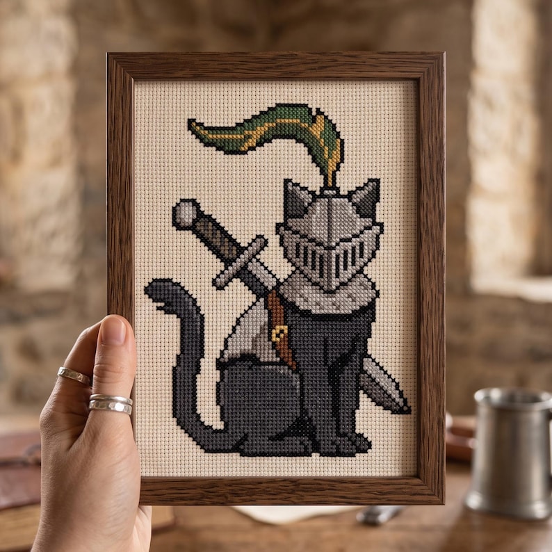 Op de afbeelding: Een ingelijst kruissteek borduurwerk met een zwarte kat die een zilveren ridderhelm draagt met een groene en gele pluim. De kat heeft een zwaard op de rug gebonden met een bruine leren riem, afgebeeld op een cr&egrave;mekleurige stoffen ondergrond.