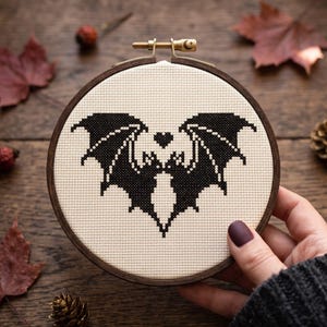 以下が含まれることがあります： クリーム色のアイーダ布が入った円形の木製刺繍枠。布には、翼を大きく広げたコウモリと、その耳の間に配置された小さなハートの形が黒いクロスステッチで刺繍されています。