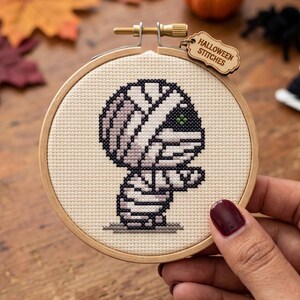 May include: A circular wooden embroidery hoop containing a cross-stitch design of a mummy wrapped in light gray bandages with a single visible green eye. A small wooden tag attached to the top of the hoop features the text Halloween Stitches.