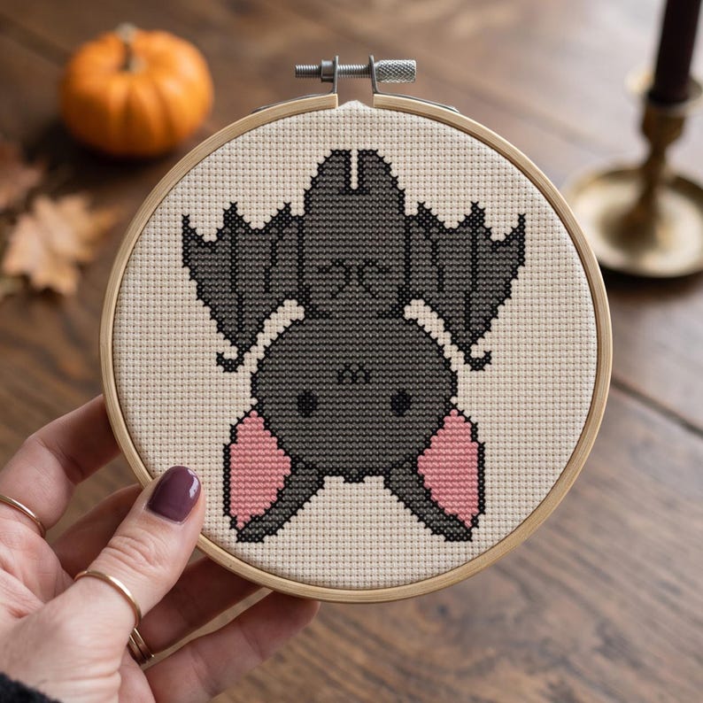 May include: A circular wooden embroidery hoop containing a cross-stitch pattern of a hanging bat. The bat is dark gray with light pink inner ears, depicted upside down against a cream-colored fabric background. The hoop features a metal screw.