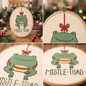 May include: An oval wooden embroidery hoop containing a cross-stitch pattern of a green frog with a yellow belly. A red bow hangs above the frog, and the text MISTLE-TOAD is stitched in black below the frog on a cream-colored fabric background.