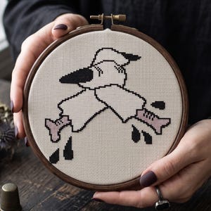 May include: A circular wooden embroidery hoop containing a cross-stitch design on cream-coloured Aida fabric. The pattern features a stylised white ghost figure with black outlines, wearing a hat, with light pink boots and scattered black droplets.