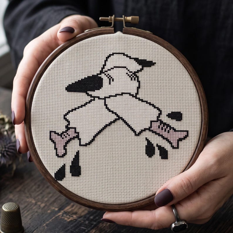 May include: A circular wooden embroidery hoop containing a cross-stitch design on cream-coloured Aida fabric. The pattern features a stylised white ghost figure with black outlines, wearing a hat, with light pink boots and scattered black droplets.