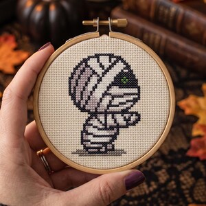 May include: A circular wooden embroidery hoop containing a cross-stitch design of a mummy character. The mummy is rendered in shades of white, light gray, and black thread on a cream-colored fabric, featuring a single visible green eye.