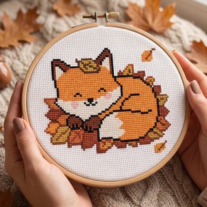 May include: A cross-stitch embroidery project in a circular wooden hoop featuring a stylized orange fox with a white chest and tail tip, curled up amidst a pile of brown and yellow autumn leaves on a white fabric background with a metal screw.