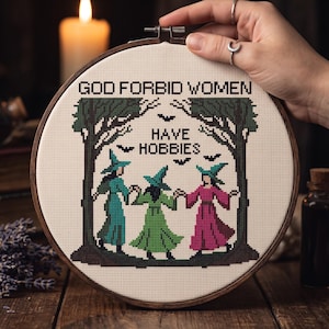 Pu&ograve; includere: Un telaio da ricamo circolare in legno contenente un disegno a punto croce su tessuto color crema. Il testo recita GOD FORBID WOMEN HAVE HOBBIES. Il disegno raffigura tre figure con cappelli a punta e abiti lunghi color ottanio, verde e rosa, in piedi tra due alberi verde scuro con piccoli pipistrelli neri che volano sopra.