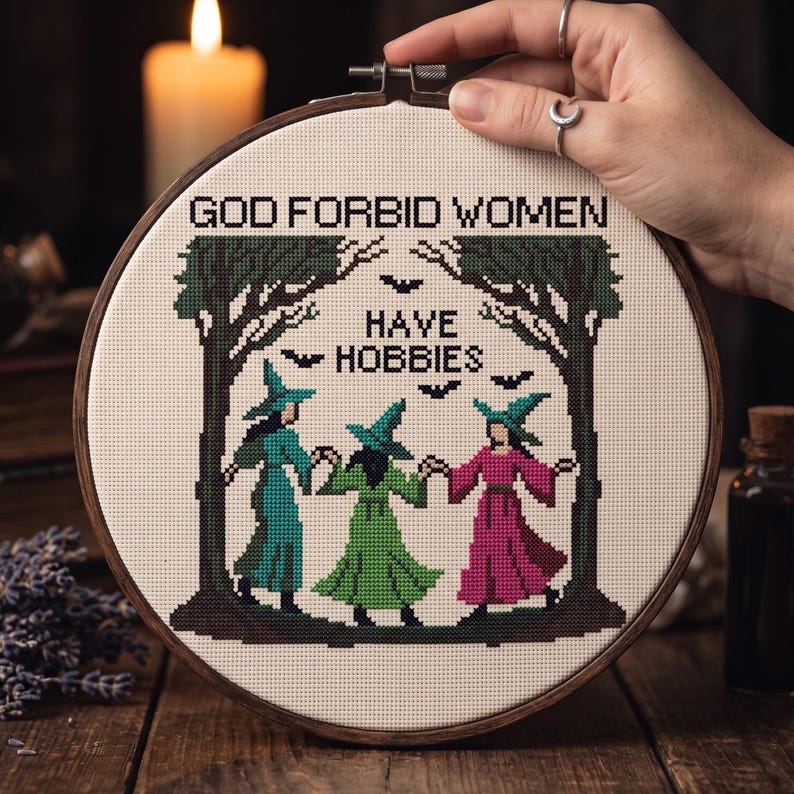 Pu&ograve; includere: Un telaio da ricamo circolare in legno contenente un disegno a punto croce su tessuto color crema. Il testo recita GOD FORBID WOMEN HAVE HOBBIES. Il disegno raffigura tre figure con cappelli a punta e abiti lunghi color ottanio, verde e rosa, in piedi tra due alberi verde scuro con piccoli pipistrelli neri che volano sopra.