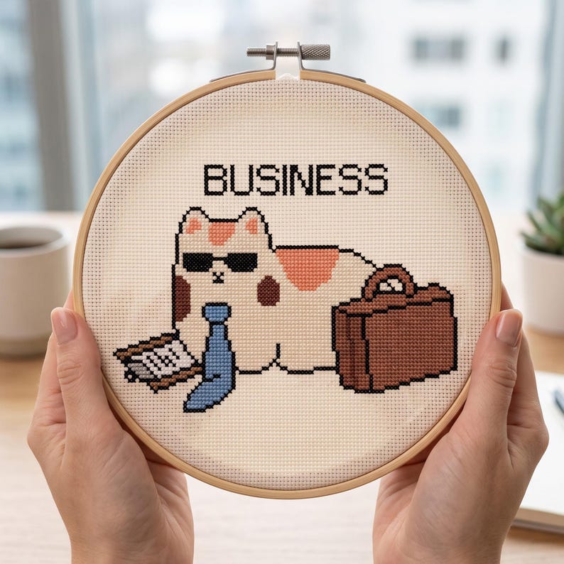 May include: A cross-stitch embroidery in a round wooden hoop featuring a calico cat wearing black sunglasses and a blue necktie. The cat is positioned next to a brown briefcase and a clipboard. The word BUSINESS is stitched in black above the cat.