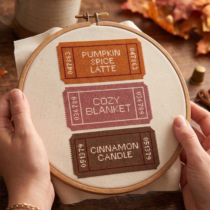 May include: A round wooden embroidery hoop containing white fabric with three stitched rectangular ticket designs. The top ticket is orange with the text PUMPKIN SPICE LATTE, the middle is mauve with COZY BLANKET, and the bottom is brown with CINNAMON CANDLE.