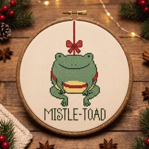 May include: A circular wooden embroidery hoop containing a cross-stitch design of a green frog hanging from a red bow. The frog has a yellow belly and is positioned above the text MISTLE-TOAD, which is stitched in dark green capital letters.