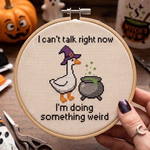 May include: A circular wooden embroidery hoop containing a cross-stitch design on cream fabric. The design features a white goose wearing a purple witch hat standing next to a grey cauldron with green liquid. Text reads: I can't talk right now I'm doing something weird.