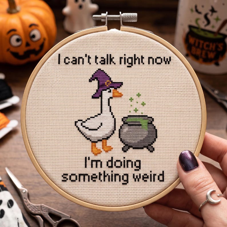 May include: A circular wooden embroidery hoop containing a cross-stitch design on cream fabric. The design features a white goose wearing a purple witch hat standing next to a grey cauldron with green liquid. Text reads: I can't talk right now I'm doing something weird.