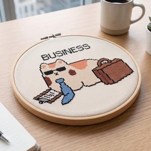 May include: A circular wooden embroidery hoop containing a cross-stitch design of a calico cat wearing black sunglasses and a blue tie. The cat holds a clipboard and a brown briefcase, with the word BUSINESS stitched in black above the cat.