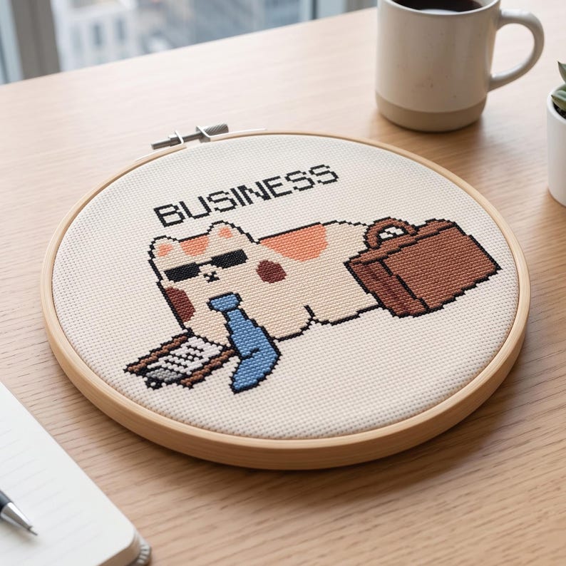 May include: A circular wooden embroidery hoop containing a cross-stitch design of a calico cat wearing black sunglasses and a blue tie. The cat holds a clipboard and a brown briefcase, with the word BUSINESS stitched in black above the cat.