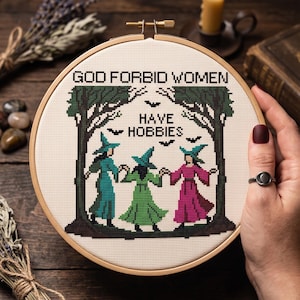 Pu&ograve; includere: Un telaio da ricamo circolare in legno contenente un disegno a punto croce di tre figure con cappelli a punta che si tengono per mano tra due alberi verde scuro. Il testo recita GOD FORBID WOMEN HAVE HOBBIES, con piccole sagome di pipistrelli.