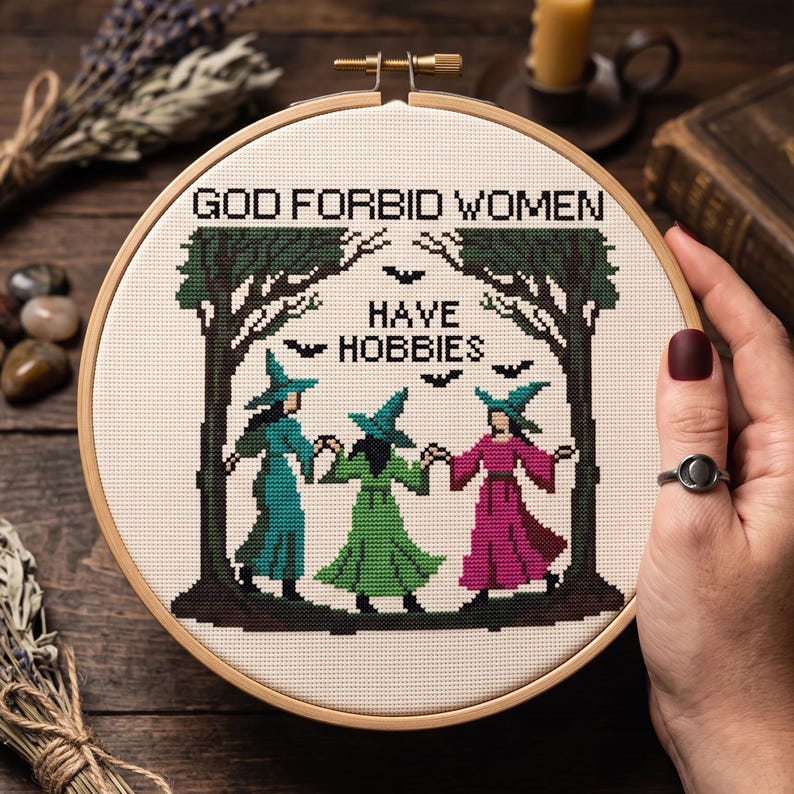 Pu&ograve; includere: Un telaio da ricamo circolare in legno contenente un disegno a punto croce di tre figure con cappelli a punta che si tengono per mano tra due alberi verde scuro. Il testo recita GOD FORBID WOMEN HAVE HOBBIES, con piccole sagome di pipistrelli.