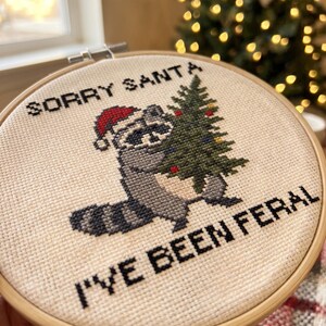 May include: A circular wooden embroidery hoop containing a cross-stitch design on cream fabric. The design features a grey raccoon wearing a red Santa hat and holding a green Christmas tree, with the black text SORRY SANTA I'VE BEEN FERAL.