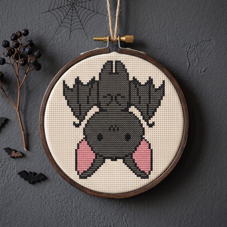 May include: A cross-stitch embroidery of a hanging bat centered in a circular wooden hoop. The bat is dark gray with light pink inner ears, stitched onto a cream-colored fabric. The hoop is suspended by a piece of twine at the top.