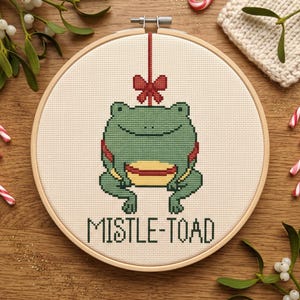 May include: A circular wooden embroidery hoop containing a cross-stitch design of a green frog with a yellow belly, positioned beneath a red bow. The text MISTLE-TOAD is stitched in dark green capital letters at the bottom of the white fabric.