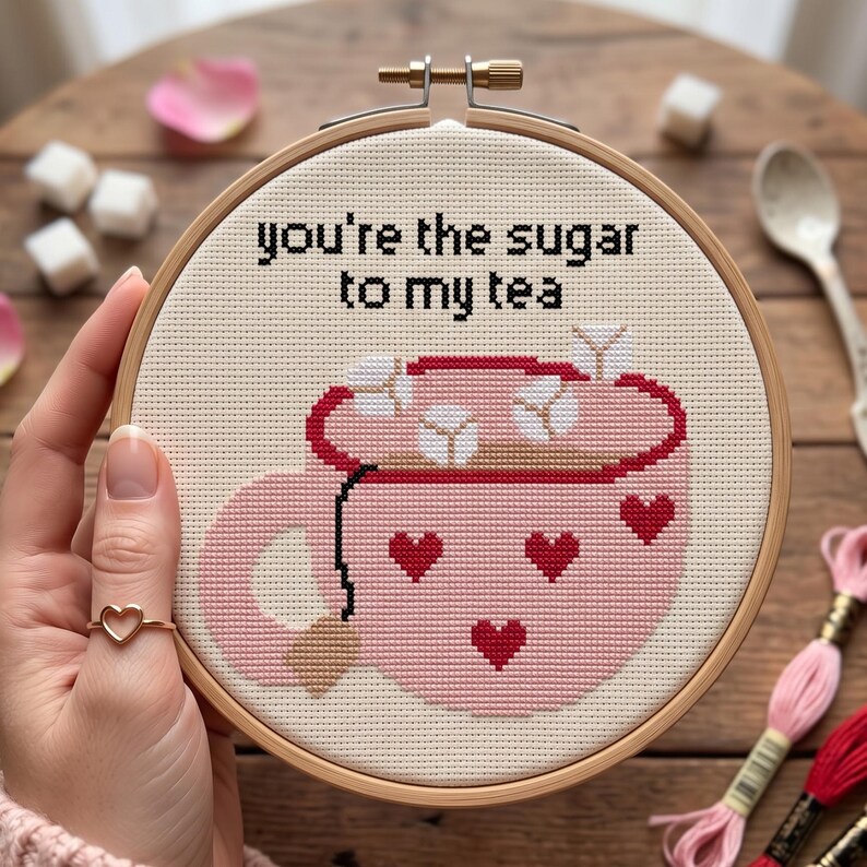 May include: A cross-stitch embroidery in a circular wooden hoop featuring a pink mug with red hearts and white sugar cubes. The text reads, you're the sugar to my tea, stitched in black thread on a cream-colored fabric background.