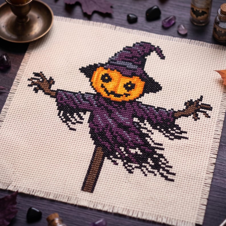 Puede incluir: Un bordado de punto de cruz sobre tela color crema que muestra un espantap&aacute;jaros con una cabeza de calabaza naranja brillante y un sombrero puntiagudo morado. El espantap&aacute;jaros viste una prenda morada harapienta con brazos de palo marrones.